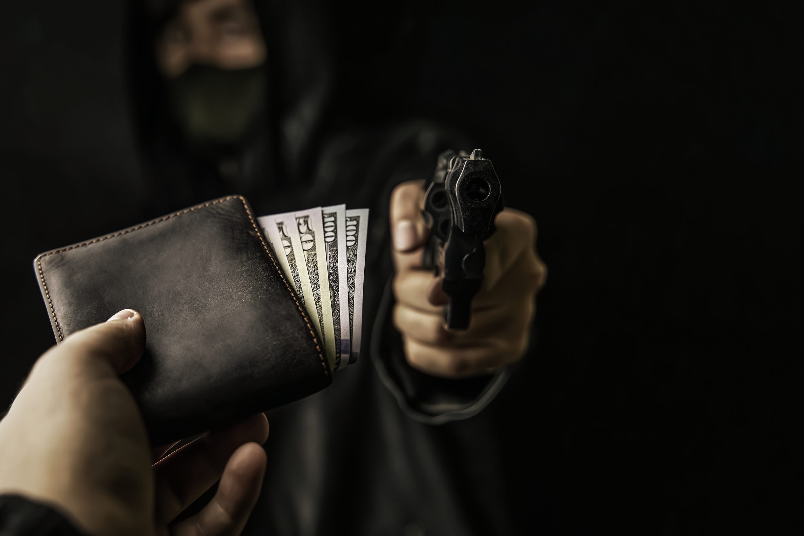 Armed robbery. Man's hand holds out purse of money to robber with gun. Firearm is pointed at camera. Criminal in hood and mask. Assault on unarmed man. Hundred and fifty dollars in open wallet.